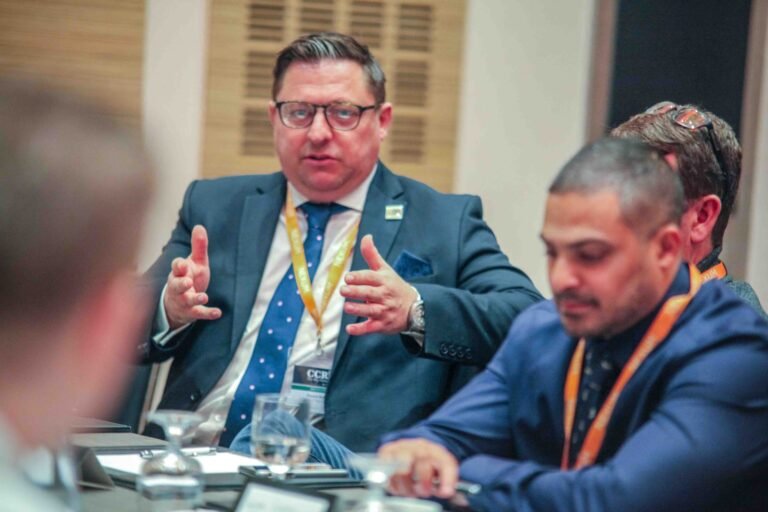 A corporate event scene features a speaker engaging with attendees at a roundtable discussion in a professional setting.